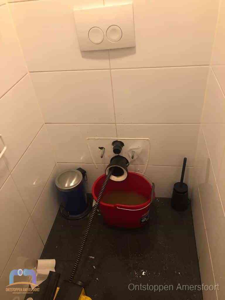 Een ontstopte wand-wc met verwijderde toiletpot waar een ontstoppingsveer in de wanduitgang verdwijnt, het achterhalen waar de verstopping zit in Amersfoort
