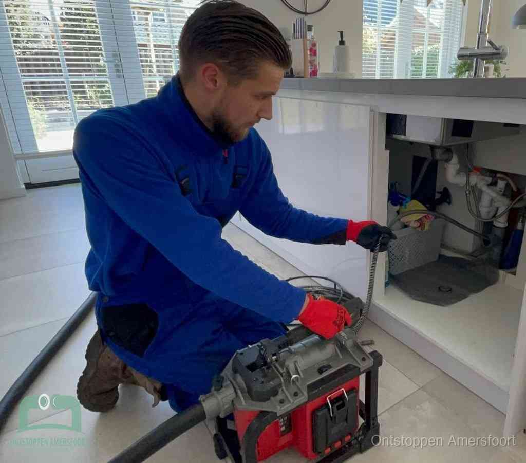 Een monteur met rode handschoenen knielt in een keuken en voert een elektrische ontstoppingsveer in tijdens het professioneel afvoer ontstoppen in Amersfoort